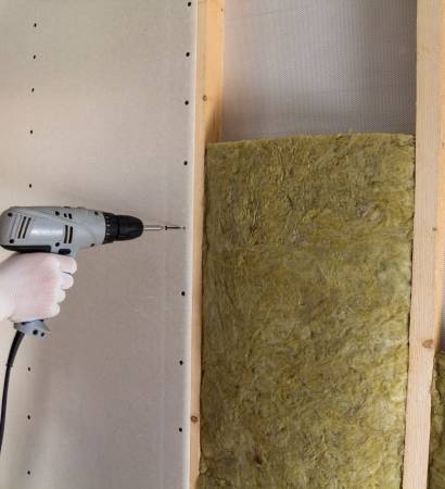 Close-up of worker hand with electric screwdriver fastening drywall to wooden timber frame with insulation mineral rock wool staff. Warm home, low energy bills, construction and renovation concept.